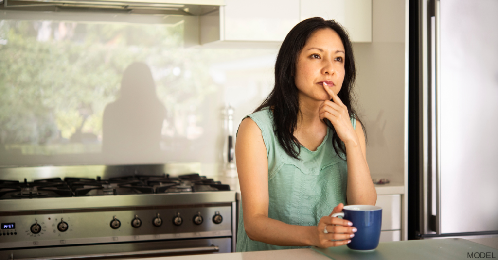A woman standing in the kitchen holding a coffee mug, who appears to be in deep thought.