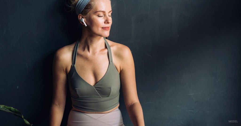 Woman wearing a green top (model) stands against a dark background with earbuds in.